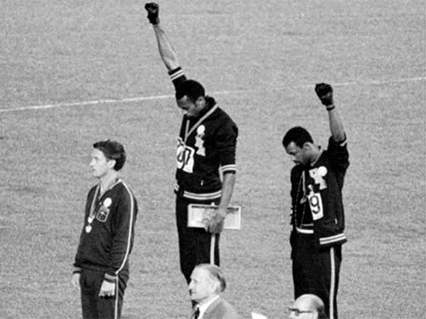 O medalhista de ouro Tommie Smith (centro) e o medalhista de bronze John Carlos (à direita) mostrando o punho levantado no pódio após a corrida de 200 m nos Jogos Olímpicos de Verão de 1968